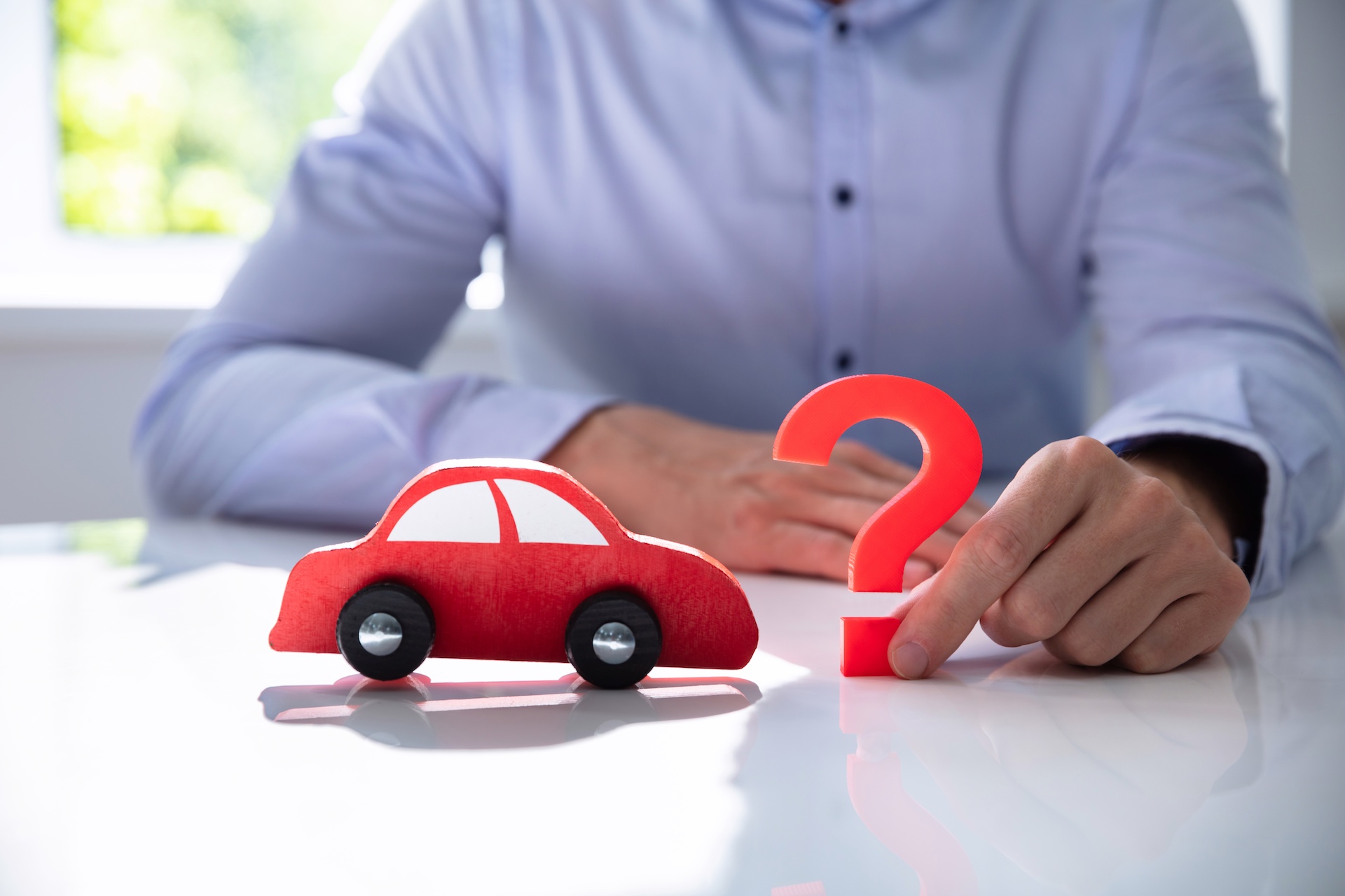 Une personne en chemise touche un point d'interrogation rouge à côté d'une petite voiture rouge en bois, symbolisant la réflexion sur le choix d'une assurance auto.