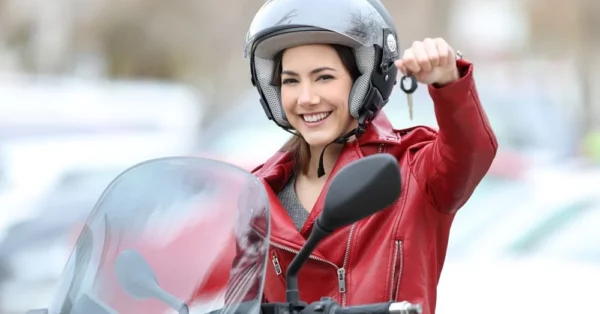 Jeune femme souriante en veste rouge et casque de moto, tenant les clés de son scooter en main