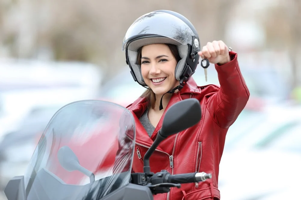 Jeune femme souriante en veste rouge et casque de moto, tenant les clés de son scooter en main