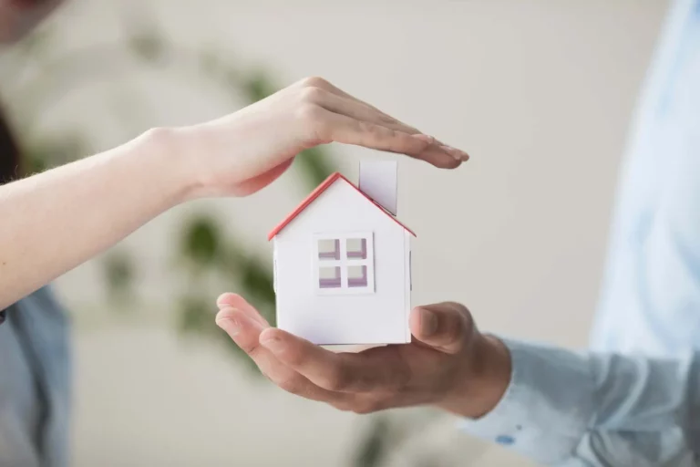 Une maison miniature protégée par des mains symbolisant la sécurité et la couverture d'une assurance emprunteur