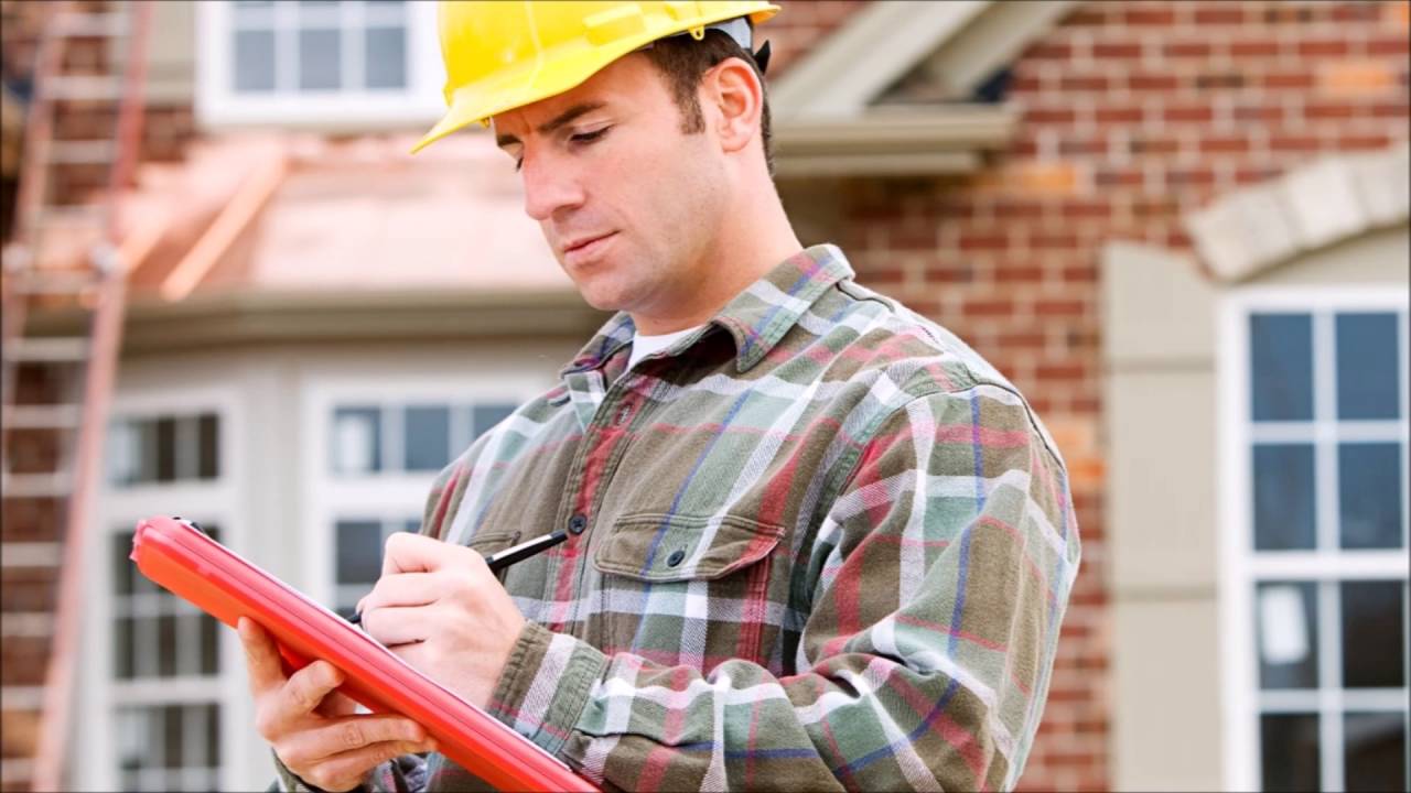 Un contre-expert en assurance, portant un casque de sécurité, prend des notes sur un bloc-notes rouge devant une maison en briques, probablement en train d'effectuer une évaluation des dommages pour une réclamation d'assurance habitation.