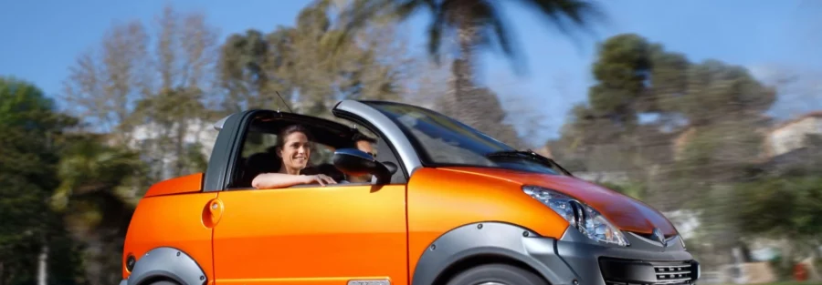 Voiturette orange en pleine conduite, avec une conductrice souriante, sous un ciel bleu et des palmiers en arrière-plan