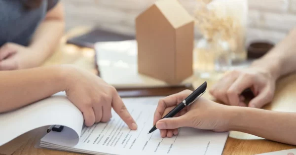 Des personnes examinent et signent un contrat d'assurance emprunteur, symbolisant une discussion sur le changement d'assurance pour un prêt immobilier, avec une petite maquette de maison en arrière-plan