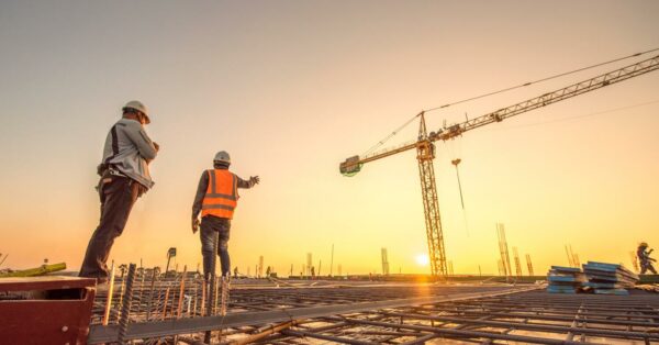 Deux ouvriers en tenue de sécurité sur un chantier de construction, discutant au coucher de soleil, avec une grue en arrière-plan.