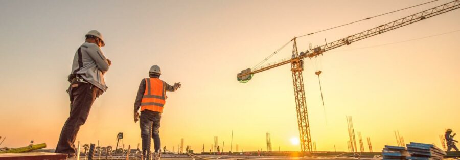 Deux ouvriers en tenue de sécurité sur un chantier de construction, discutant au coucher de soleil, avec une grue en arrière-plan.