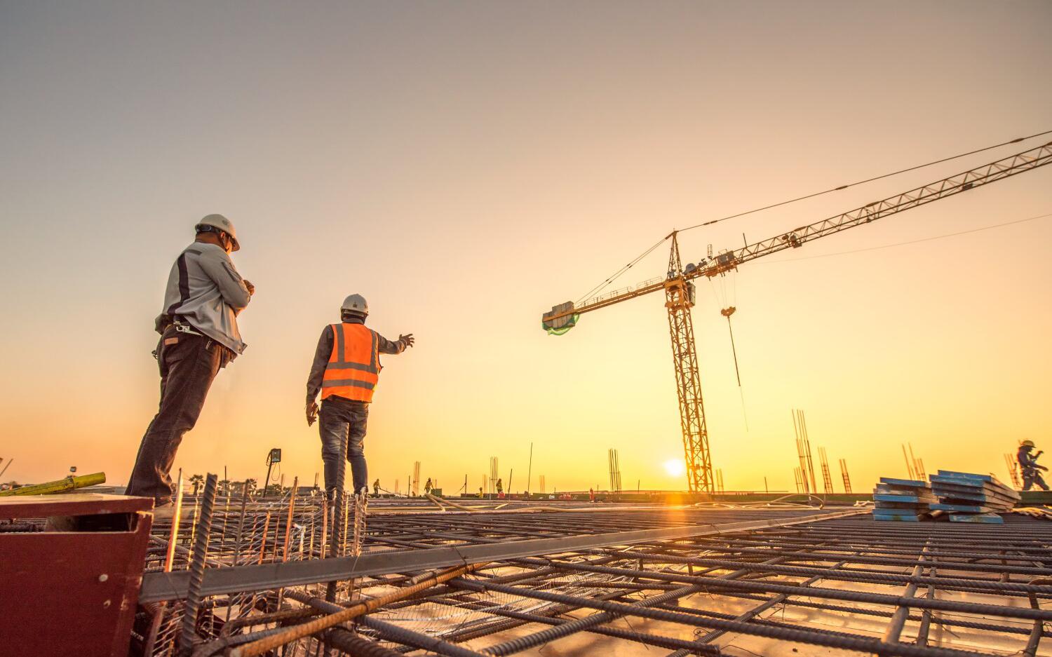 Deux ouvriers en tenue de sécurité sur un chantier de construction, discutant au coucher de soleil, avec une grue en arrière-plan.