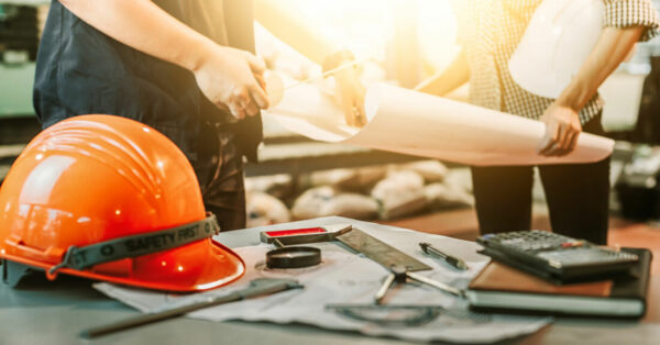 Casque de chantier orange et outils de construction posés sur une table, avec deux professionnels examinant des plans en arrière-plan, symbolisant la planification et la sécurité dans les travaux de construction.