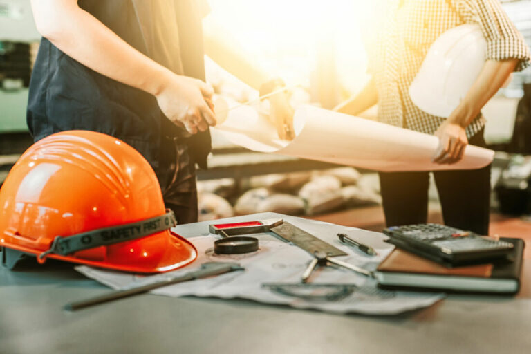 Casque de chantier orange et outils de construction posés sur une table, avec deux professionnels examinant des plans en arrière-plan, symbolisant la planification et la sécurité dans les travaux de construction.