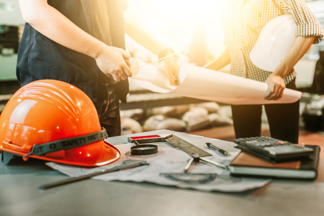 Casque de chantier orange et outils de construction posés sur une table, avec deux professionnels examinant des plans en arrière-plan, symbolisant la planification et la sécurité dans les travaux de construction.